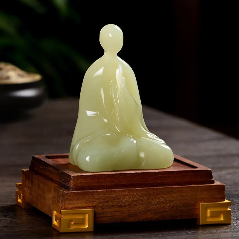 A pale yellow-green jade-like seated Buddha statue displayed on a dark wooden base with gold-tone corner accents, set against a dark blurred background.