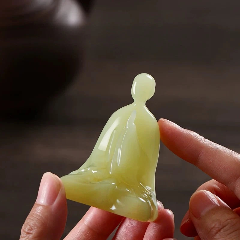 A small polished jadeite figurine of a seated Buddha, held between two fingers to show its translucency and color against a dark, blurred background.
