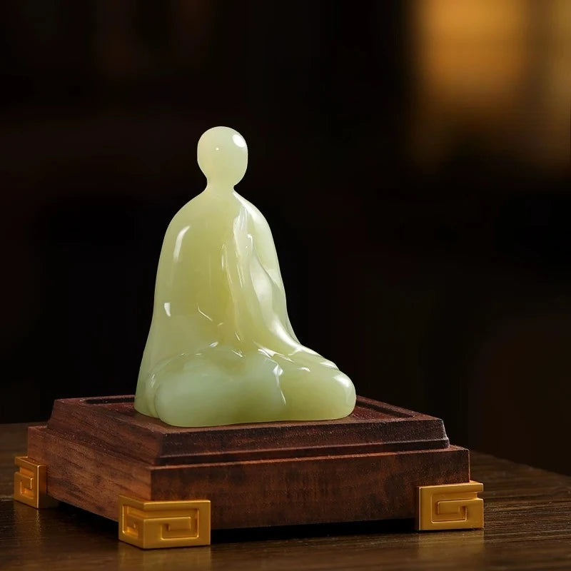 A pale green jade-like seated Buddha statue displayed on a dark wooden pedestal with gold-toned corner accents, set against a dark blurred background.