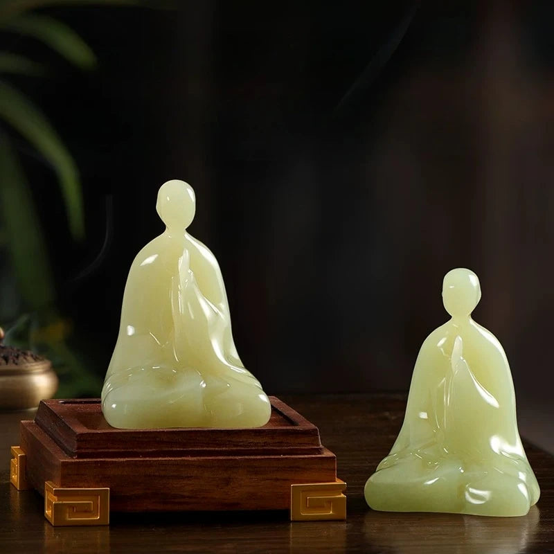Two pale yellow-green jade-like seated figurines displayed on a dark wooden surface against a dark blurred background. One figurine is larger and placed on a rectangular wooden base with gold-toned corner accents, while the other is smaller standing beside it.