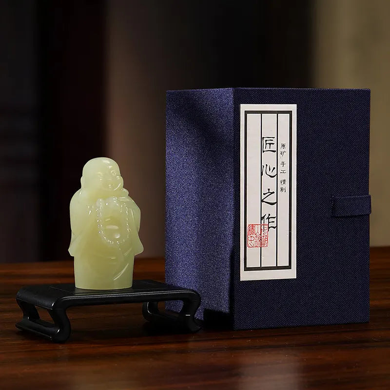 A small pale green carved figurine displayed on a black stand beside a dark navy presentation box with a white label and red seal, set on a wooden surface.