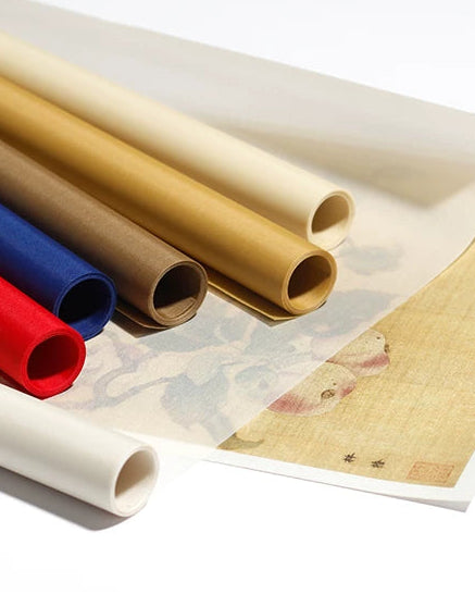 Several rolled sheets of paper or canvas in cream, tan, brown, blue, and red are arranged on a white surface, with a partially unrolled beige sheet showing a faint printed design.