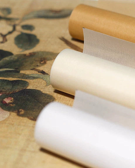 Three rolls of textured paper or fabric in white, cream, and tan, arranged diagonally on a beige surface with a leafy pattern.