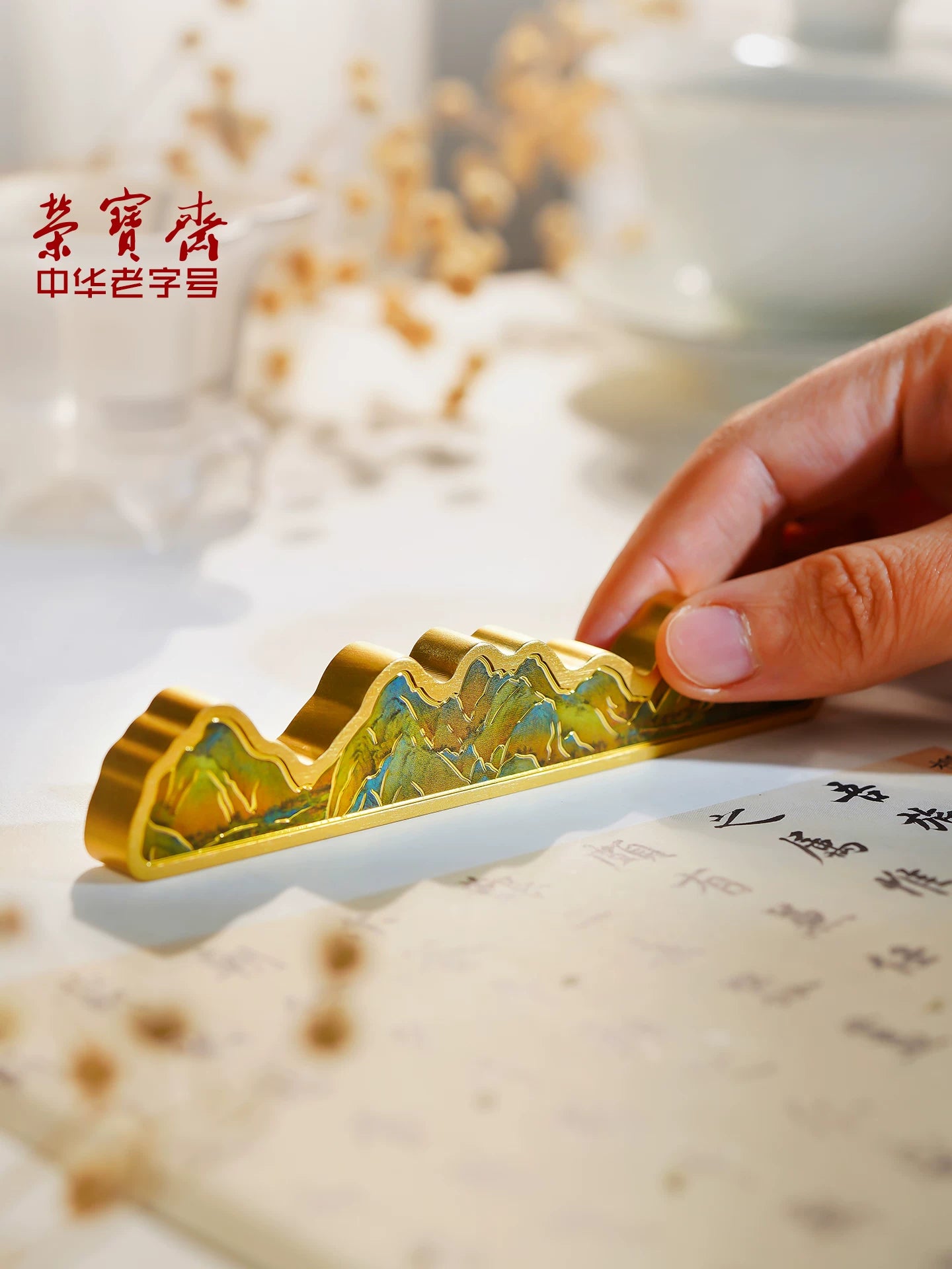 Hand holding Rongbaozhai brass brush rest with A Thousand Miles of Rivers and Mountains enamel design